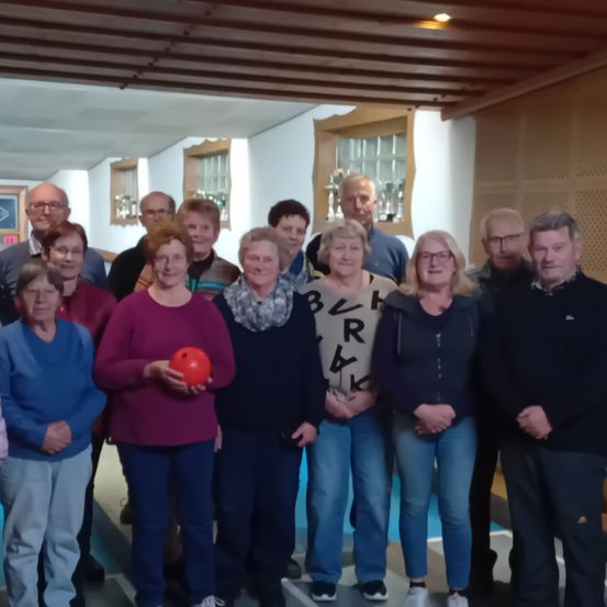 Eine Gruppe von Menschen posiert für ein Foto in einer Bowlingbahn. Sie alle lächeln und einige halten Bowlingkugeln. Der Raum hat Holzbalken und weiße Wände.