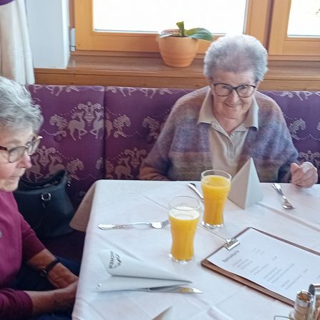 Zwei ältere Frauen sitzen an einem Tisch und sehen sich an. Sie tragen beide eine Brille und lächeln. Orangensaft, Besteck und eine Speisekarte sind auf dem Tisch. Ein Topf steht auf der Fensterbank.