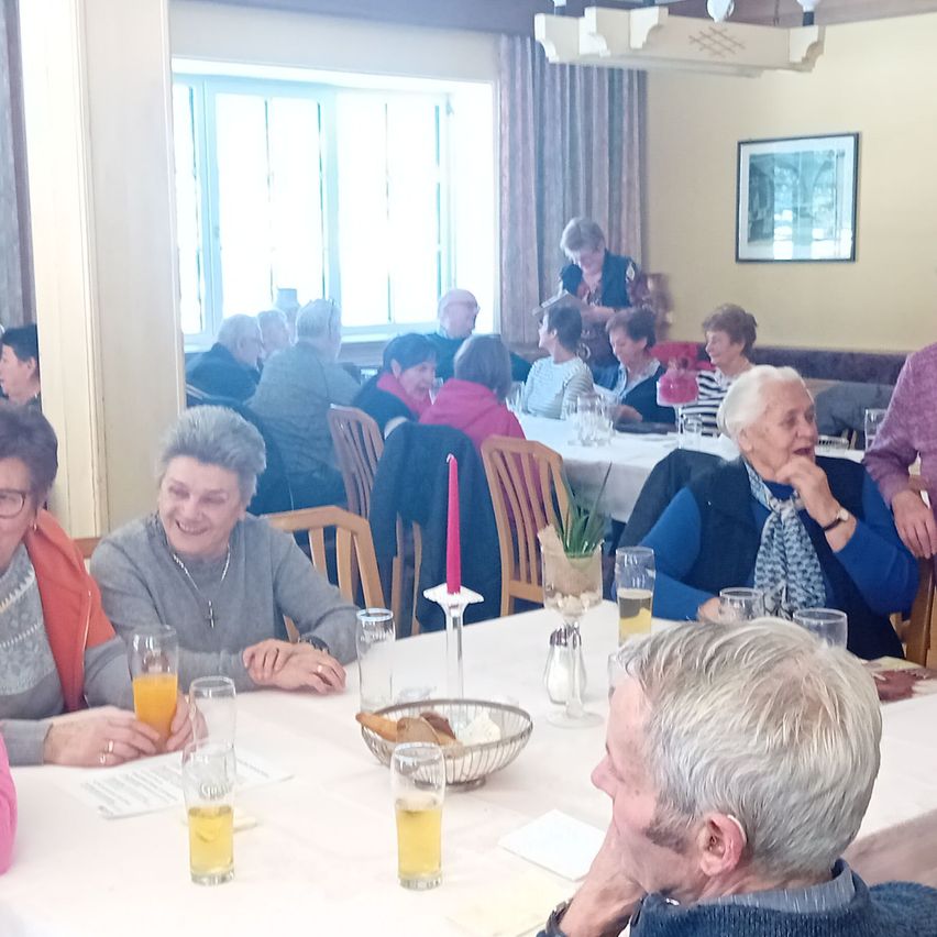 Eine Gruppe älterer Frauen sitzt an einem Tisch und trinkt Orangensaft. Gläser und eine Kerze stehen auf dem Tisch, und der Raum hat eine gelbe Wand und einen Bilderrahmen.