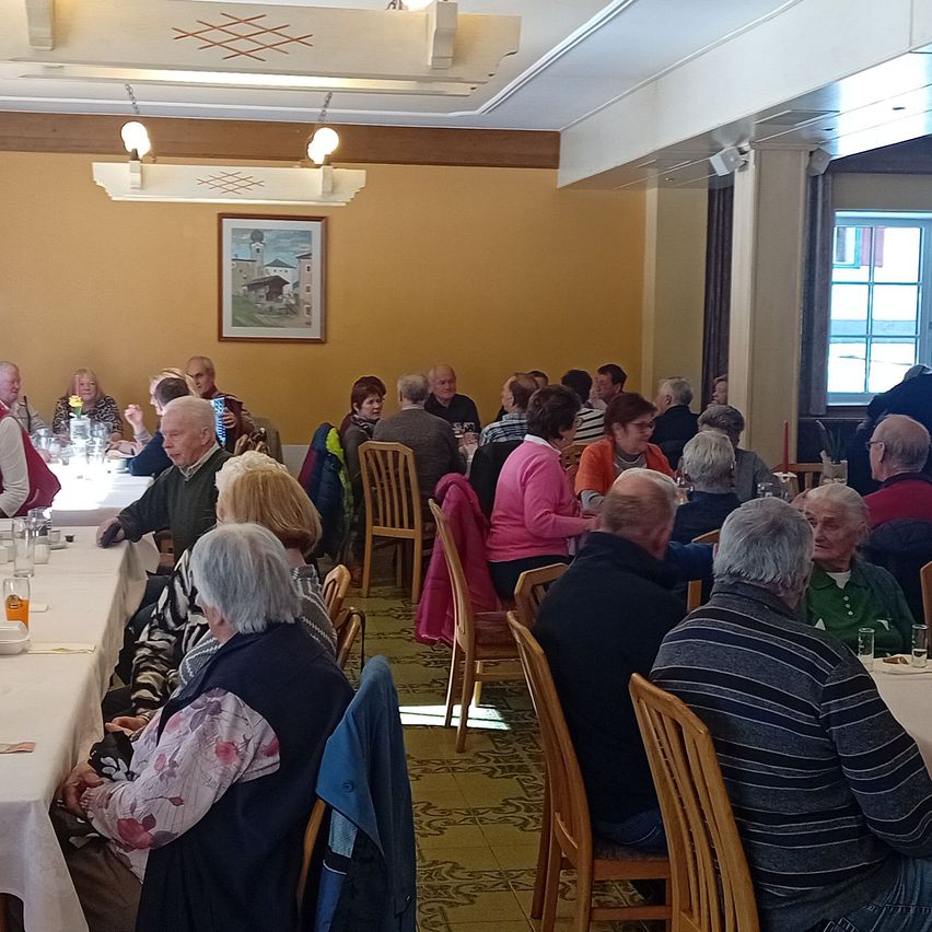 Ein Speisesaal mit älteren Menschen, die an Tischen sitzen. Einige unterhalten sich. Der Raum ist hell beleuchtet mit Lampen und hat einen Bilderrahmen an der Wand.