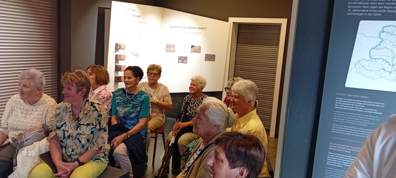 Eine Gruppe älterer Frauen sitzt in einem Raum, wahrscheinlich einem Museum, und betrachtet ein Schautafel mit fremdsprachigem Text. Eine Frau trägt eine Brille und eine Halskette.