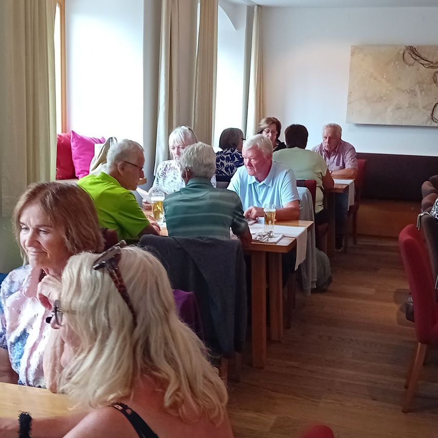 Eine Gruppe von Menschen sitzt an Tischen in einem Restaurant, einige lächeln und halten Getränke. Der Raum hat Holzböden und weiße Wände.