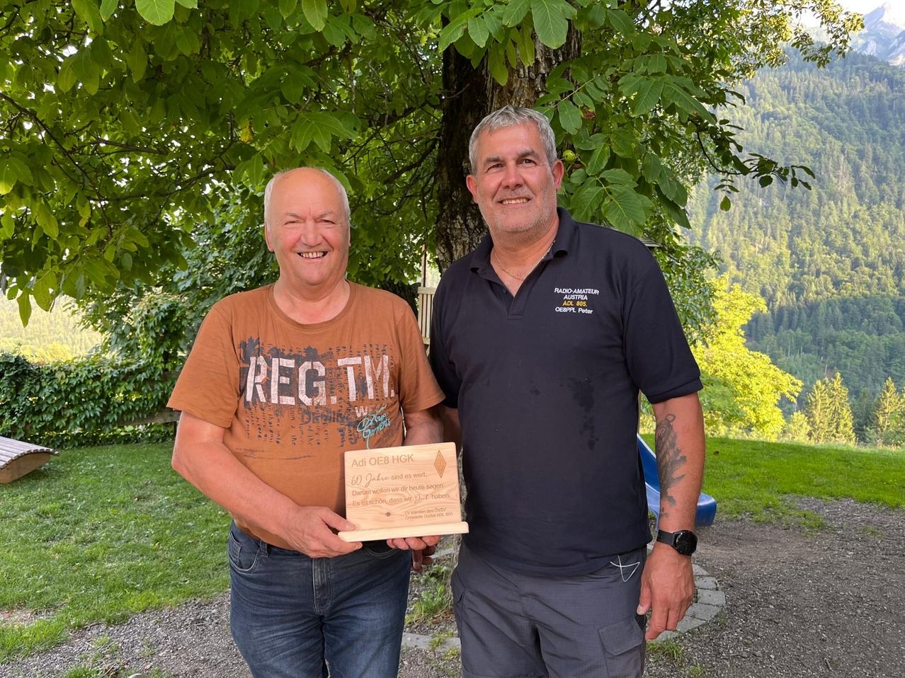 Zwei Männer lächeln unter einem Baum mit Bergen im Hintergrund. Der Mann auf der linken Seite hält eine Holzplakette. Der Mann auf der rechten Seite trägt ein blaues Polohemd mit Logo.