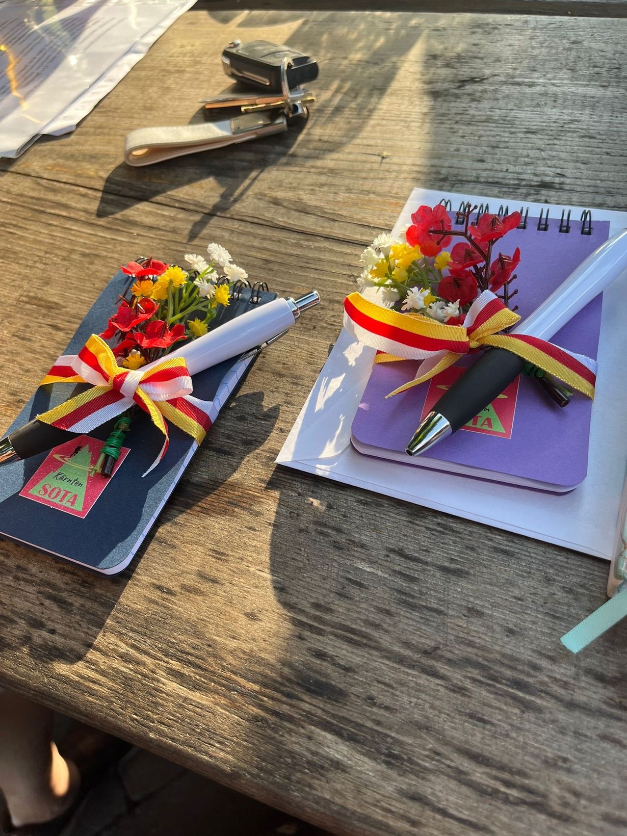 Zwei Notizbücher mit Blumen und Stiften auf einem Holztisch. Eines hat ein rotes Etikett mit der Aufschrift 'SOIA' und das andere hat einen lila Umschlag mit einer Schleife.