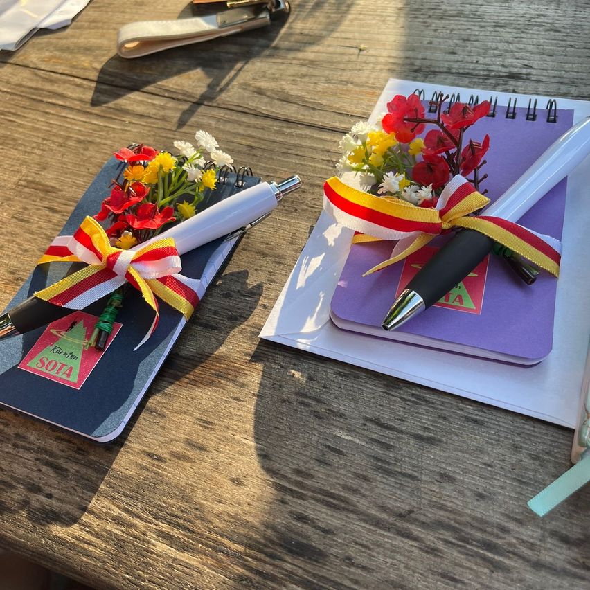 Zwei Notizbücher mit Blumen und Stiften auf einem Holztisch. Eines hat ein rotes Etikett mit der Aufschrift 'SOIA' und das andere hat einen lila Umschlag mit einer Schleife.