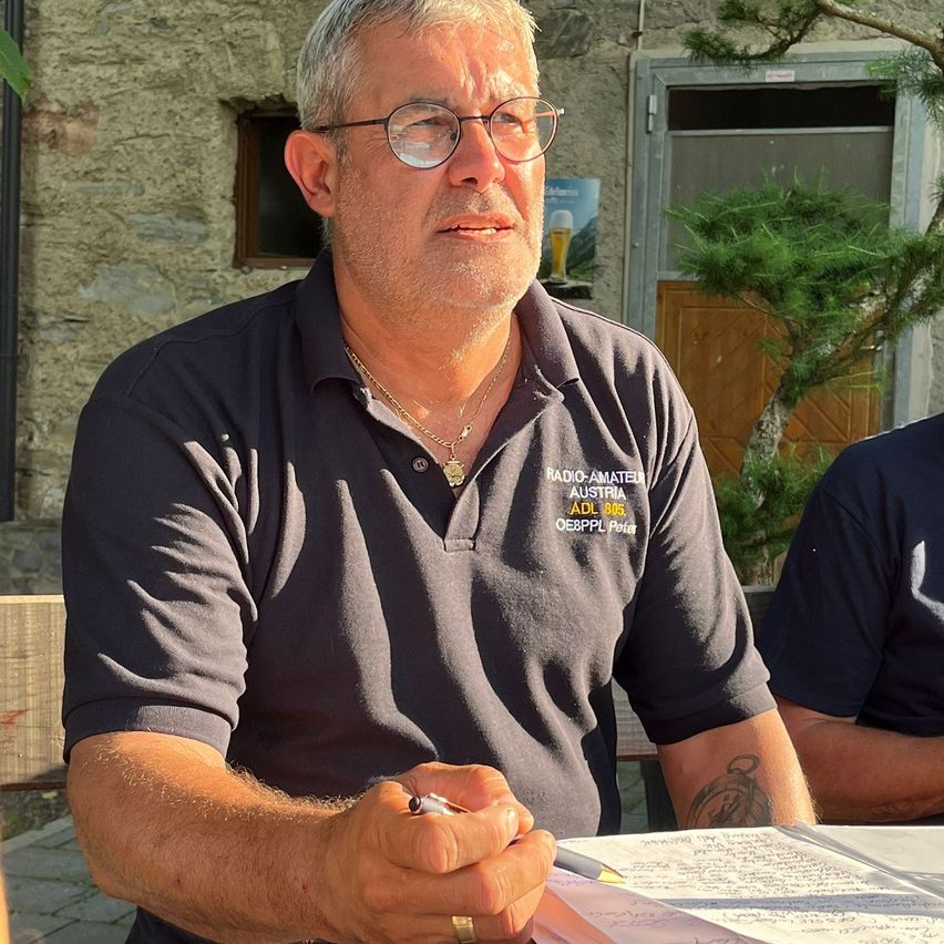 Ein Mann in einem schwarzen Hemd mit Radio Amateur Austria und ADL 006 sitzt am Tisch mit einem Stift in der Hand.