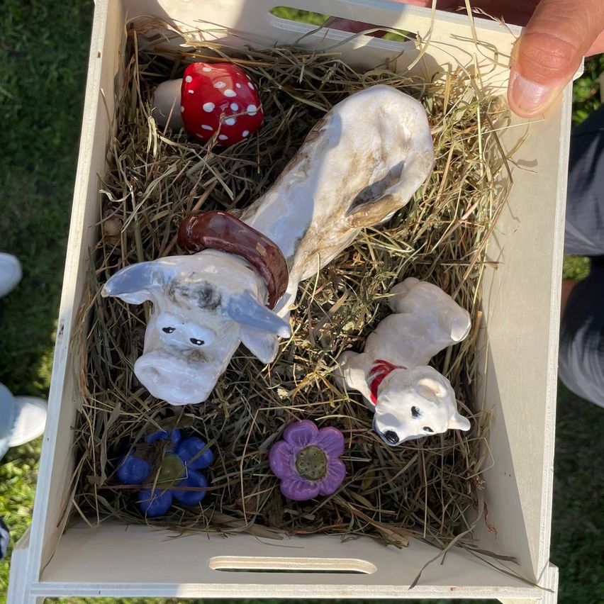 Eine Holzkiste mit Stroh enthält zwei Keramikhunde, einen mit braunem Halsband und den anderen mit rotem Halsband. In der Kiste befinden sich auch ein roter Pilz und einige Blumen.