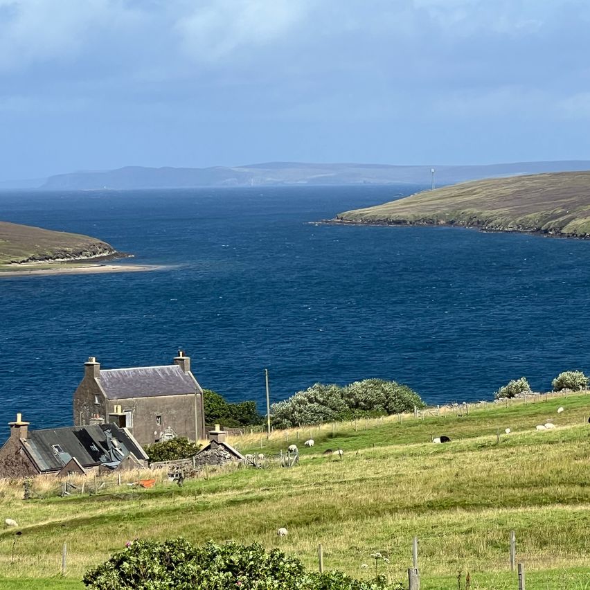 Eine Landschaft mit einem Haus am Meer, einem bewölkten Himmel und einer entfernten Bergkette.