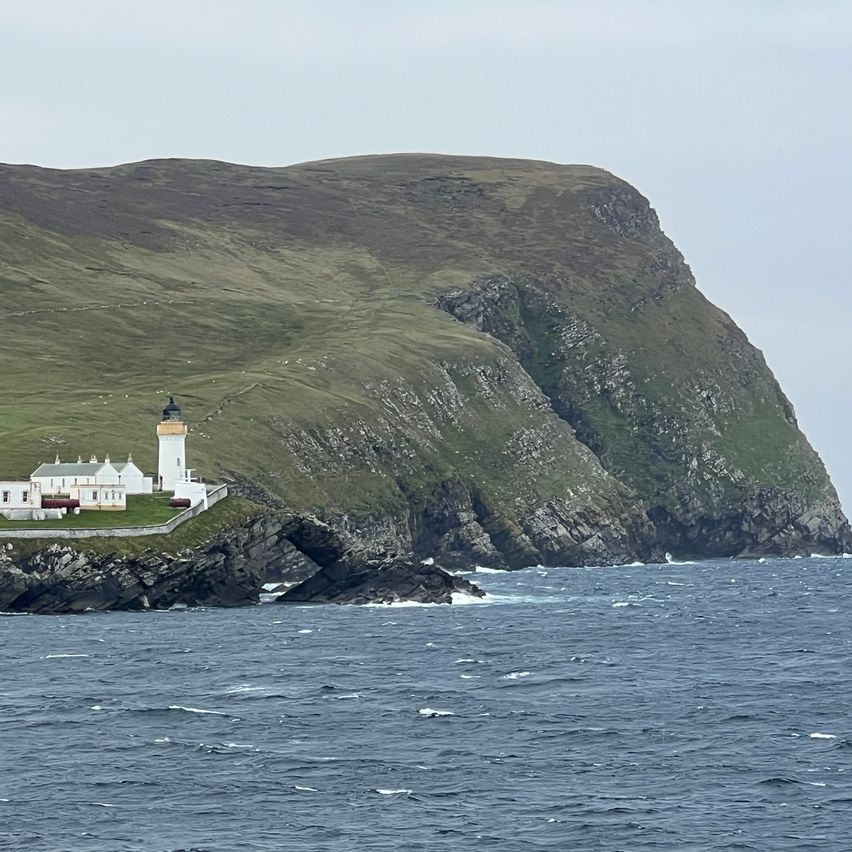 Ein weißer Leuchtturm steht auf einer Klippe am Meer, mit einem kleinen weißen Gebäude in der Nähe. Die Landschaft besteht aus grünen Hügeln und rauen Felsen. Das Meer ist ruhig mit sanften Wellen.