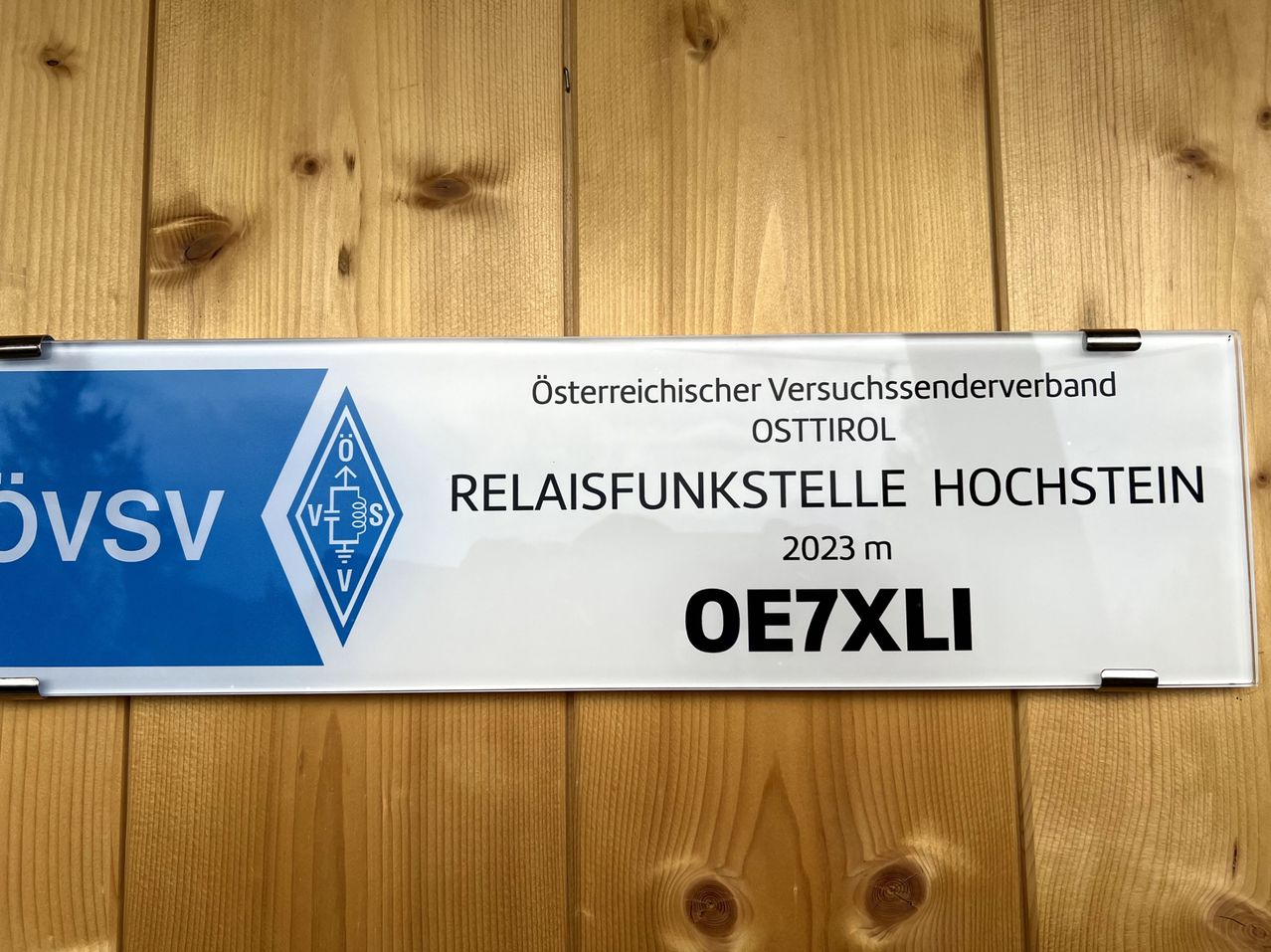 Ein weiß-blaues Schild zeigt 'Österreichischer Versuchssenderverband OSTTIROL Relaisfunkstelle HOCHST 2023 m OE7XLI' an einer Holzwand.