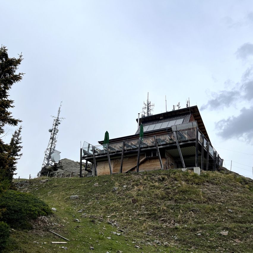 Ein Gebäude auf einem Hügel mit Balkon und Stühlen. Dahinter steht ein Turm mit Antennen.