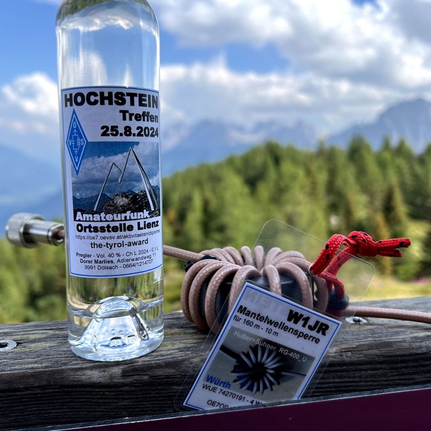 Eine Flasche mit der Aufschrift Hochstein Treffen, datiert auf 25.8.2024, steht auf einer Holzfläche mit einem Seil und einem weißen Etikett. Das Etikett trägt die Aufschrift Mantelwellensperre. Im Hintergrund sind Bäume und Berge zu sehen.