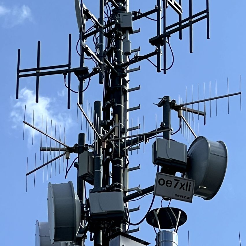 Ein hoher Telekommunikationsturm mit mehreren Antennen und Geräten, gekennzeichnet durch 'oe7xli'. Der Himmel ist klar und blau.