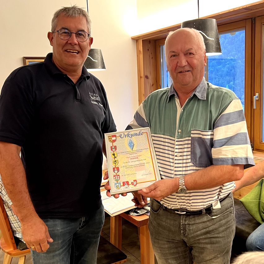 Zwei Männer stehen in einem Raum, einer hält ein Zertifikat mit mehreren Logos und Text. Der Mann auf der linken Seite trägt ein schwarzes Poloshirt mit Logo und Brille. Der Mann auf der rechten Seite trägt ein grün-weiß gestreiftes Hemd und eine Uhr.