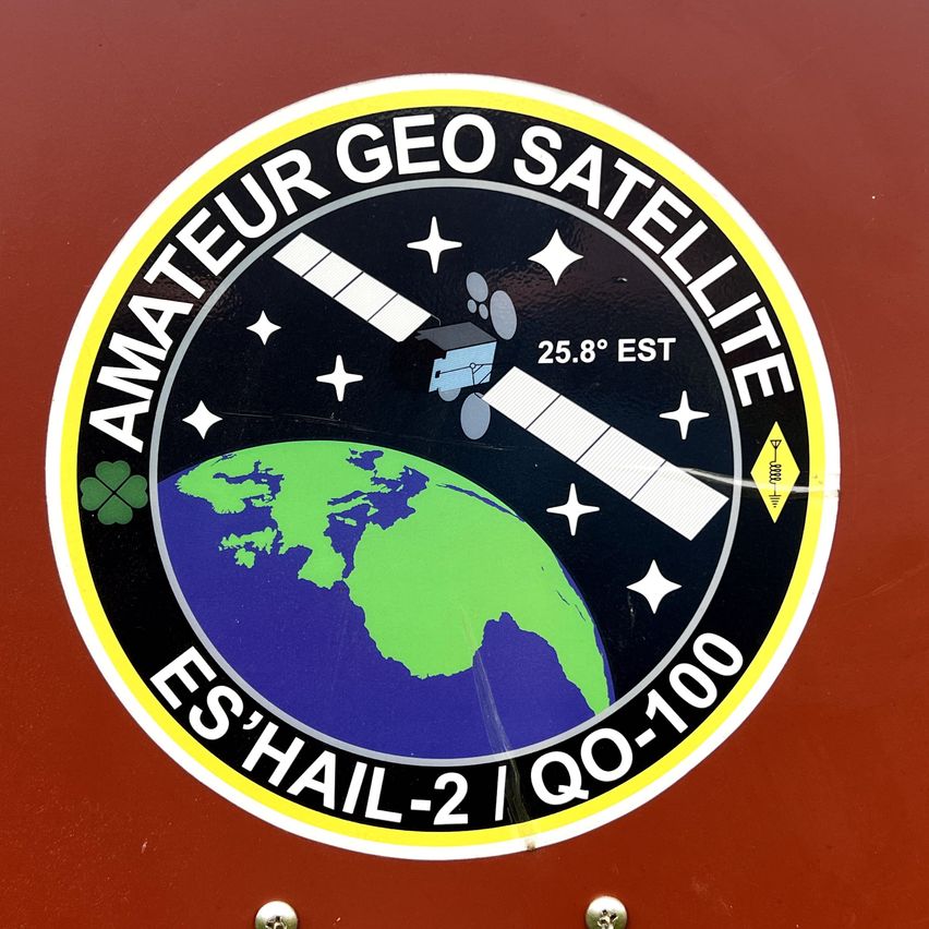 Ein rundes Abzeichen mit einem Satelliten, der die Erde umkreist, beschriftet mit 'Amateur Geo Satellit', 25,8 Grad EST und 'ES'HAIL-2 / QQ-100'.