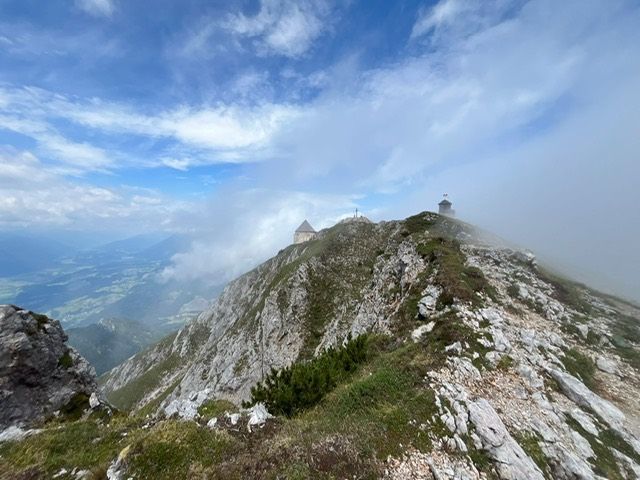 Berggipfel mit zwei kleinen Strukturen, nebliges Tal darunter, felsiges Gelände und blauer Himmel mit Wolken.