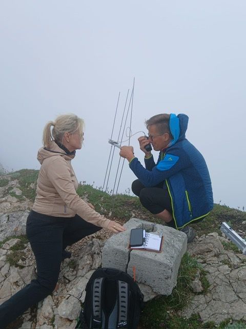 Zwei Personen befinden sich auf einem Berg, einer kauert sich hin und benutzt ein Walkie-Talkie, der andere sitzt, beide sind in Winterkleidung gekleidet.