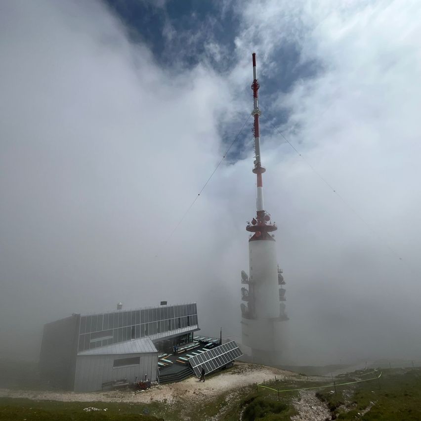 Ein großer weißer und roter Telekommunikationsturm steht auf einem nebligen Hügel neben einem Gebäude mit Solarpanelen.