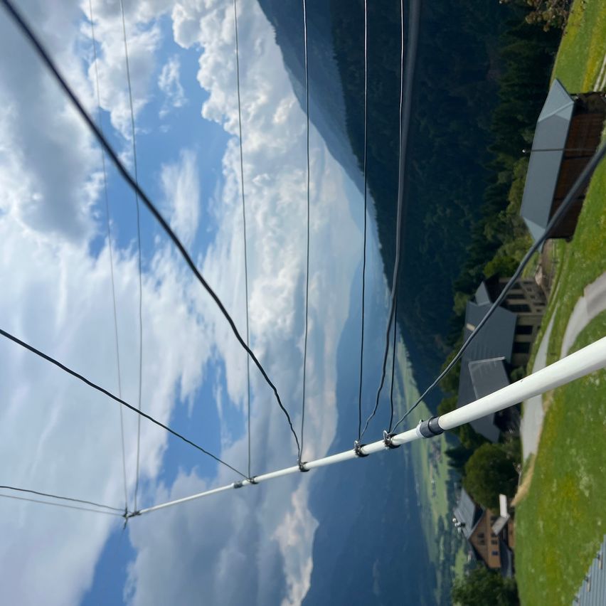 Luftaufnahme von Drähten, die mit einem hohen Mast verbunden sind, mit Hintergrund von Bergen, Wolken und Häusern.
