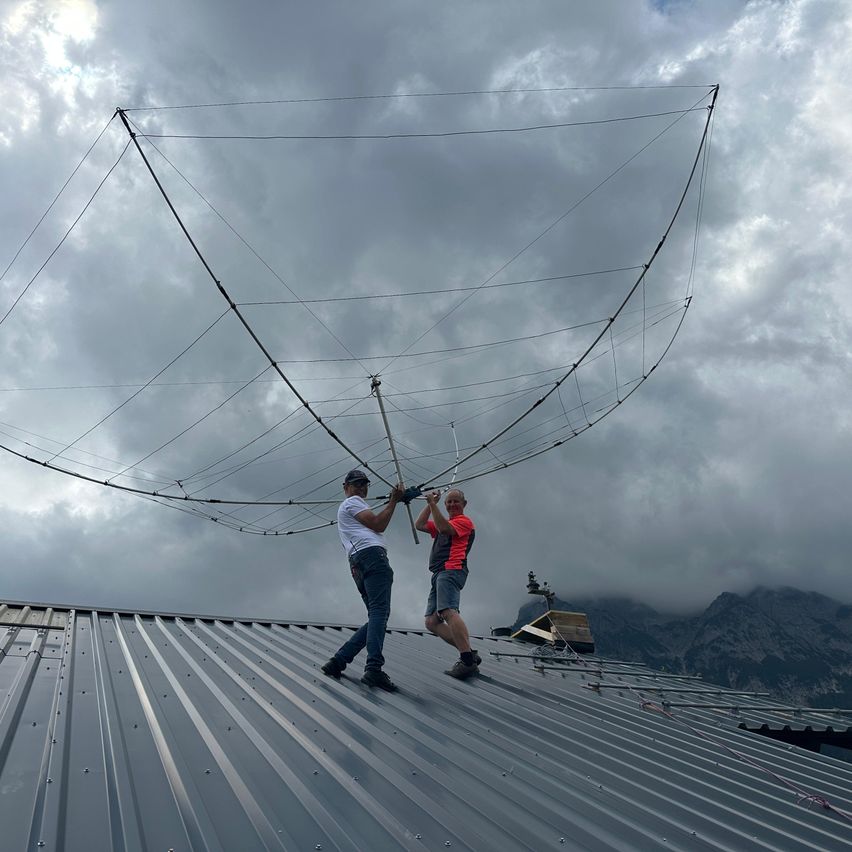 Zwei Männer arbeiten auf einem Dach bei bewölktem Himmel und stellen eine große Antennenstruktur ein. Einer trägt ein weißes T-Shirt und Jeans, der andere ein rotes und schwarzes T-Shirt und Shorts.
