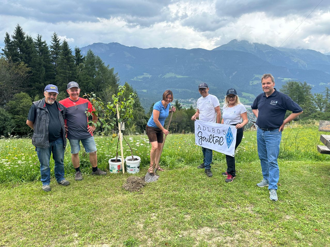 Eine Gruppe von sechs Personen steht auf einem Rasen, möglicherweise in einem Gebirgsgebiet. Sie lächeln und posieren für ein Foto. In der Mitte hält eine Frau eine Schaufel und einen Baum. Hinter ihnen ist ein Blick auf die Berge.
