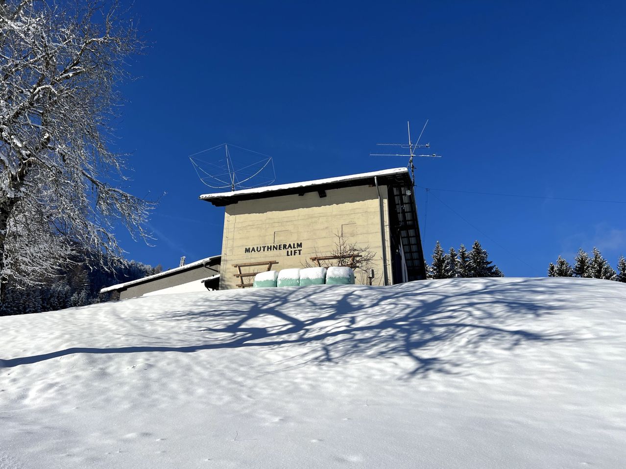 Eine Skiliftstation in Mauthnenalm, bedeckt mit Schnee, mit Bäumen und einem klaren blauen Himmel im Hintergrund.