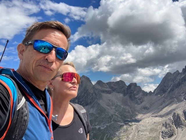 Ein Mann und eine Frau stehen auf einem Berggipfel mit Sonnenbrillen, tragen Rucksäcke und betrachten die Aussicht.