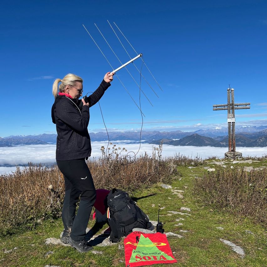Eine Frau auf einem Berggipfel hält eine Radioantenne. Sie trägt eine Jacke und Hose. In der Nähe liegen ein Rucksack und ein roter Stoff mit dem Wort 'SOTA' auf dem Boden. Im Hintergrund steht ein Kreuz auf einer grasbewachsenen Fläche mit einer entfernten Bergkette.