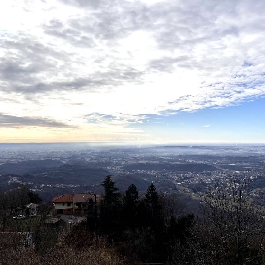 Ein malerischer Blick von einem Hügel zeigt eine Stadt mit vielen Gebäuden und einem bewölkten Himmel. Bäume umgeben die Häuser, und der Boden ist mit trockenem Gras bedeckt. Die Sonne geht am Horizont unter.