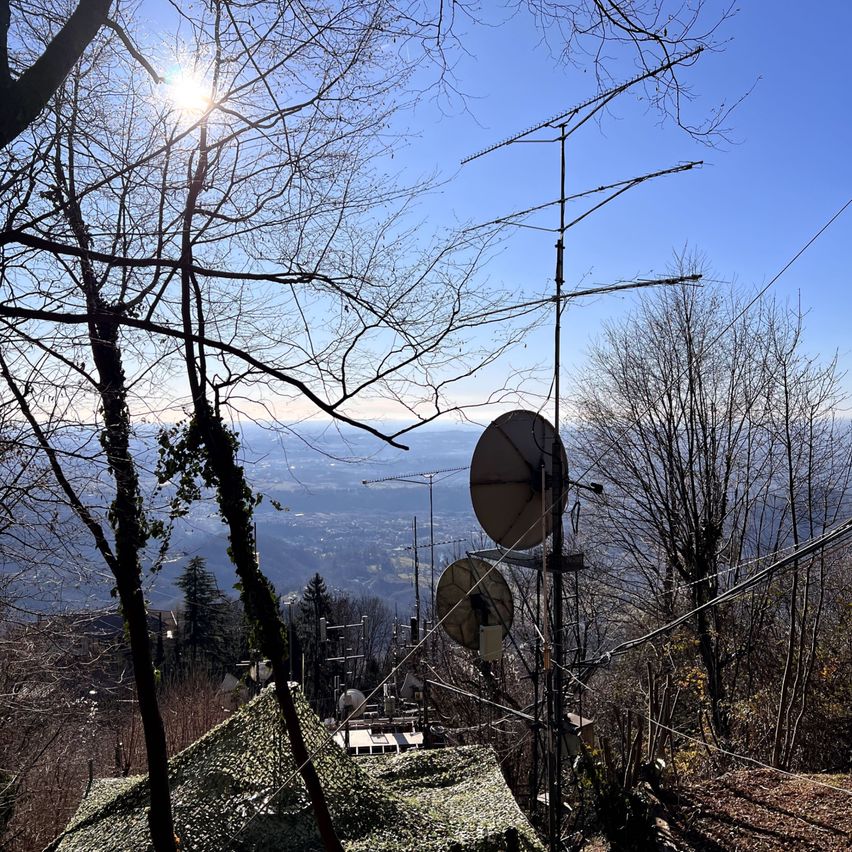 Eine Satellitenschüssel ist auf einem Mast über einem getarnten Dach montiert, umgeben von Bäumen und Antennen. Die Sonne scheint hell am klaren blauen Himmel.