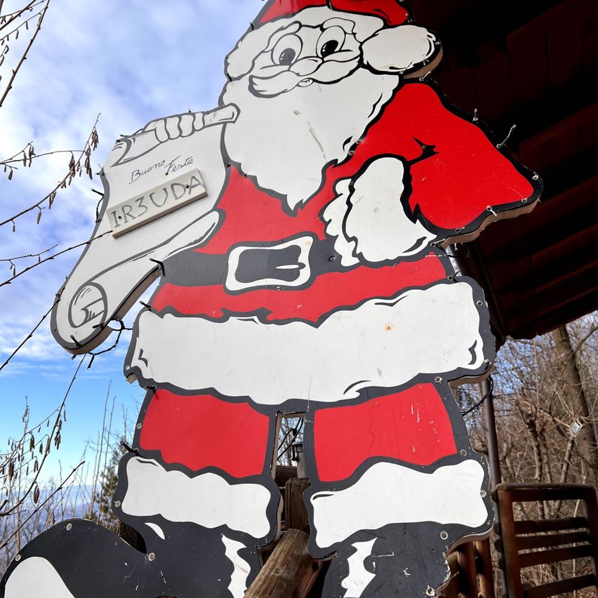 Ein großer Ausschnitt von Santa Claus, der eine Schraubenschlüssel hält, ist draußen ausgestellt. Der Ausschnitt ist rot und weiß bemalt und hat das Wort 'IR3UDA' auf einem weißen Brett. Der Hintergrund zeigt einen blauen Himmel mit einigen Wolken und einer hölzernen Struktur.