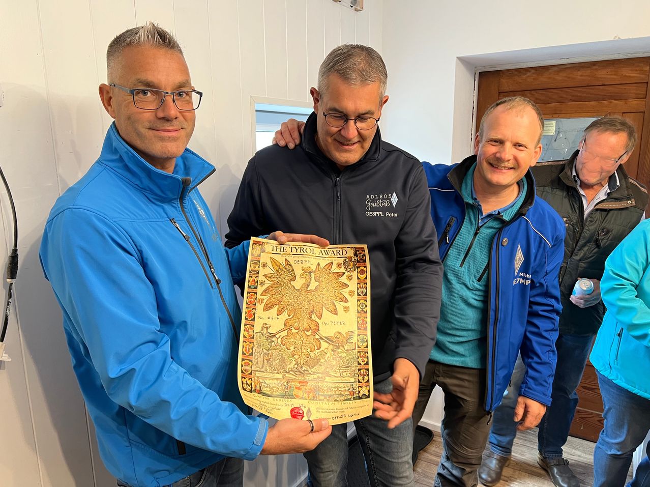 Drei Männer posieren drinnen, einer hält ein großes Zertifikat, The Tyrol Award. Alle tragen Winterjacken. Der Mann auf der linken Seite trägt eine Brille und eine blaue Jacke. Der Mann in der Mitte trägt eine schwarze Jacke und hat seinen Arm um den Mann auf der rechten Seite, der ein hellblaues Hemd und eine blaue Jacke trägt.