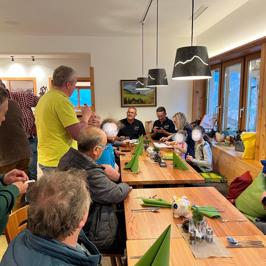 Eine Gruppe von Menschen sitzt um einen Esstisch in einem Restaurant. Einige haben Getränke und Essen auf dem Tisch, während andere Fotos machen. Ein Bilderrahmen und Lampen hängen an der Wand.