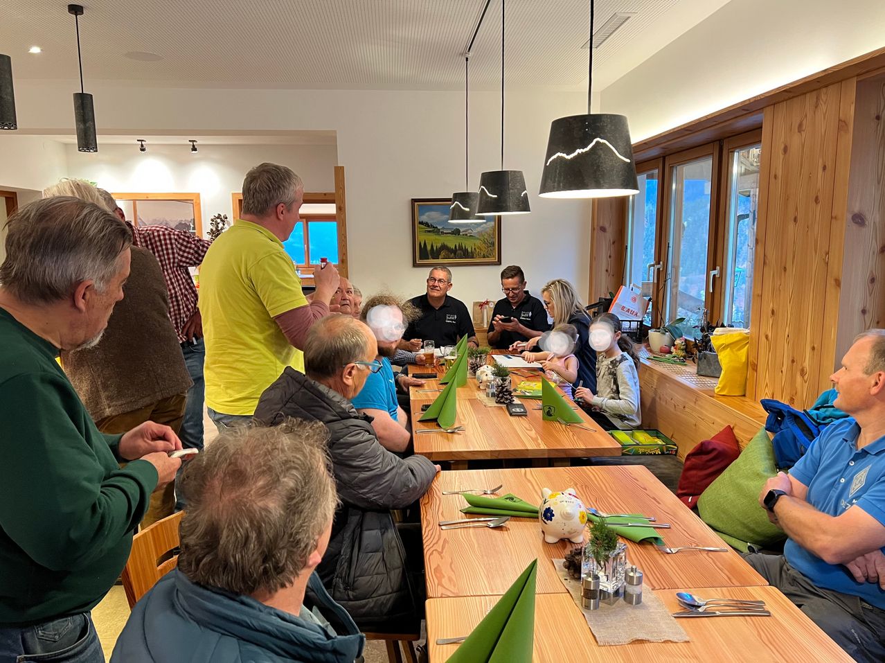 Eine Gruppe von Menschen sitzt um einen Esstisch in einem Restaurant. Einige haben Getränke und Essen auf dem Tisch, während andere Fotos machen. Ein Bilderrahmen und Lampen hängen an der Wand.