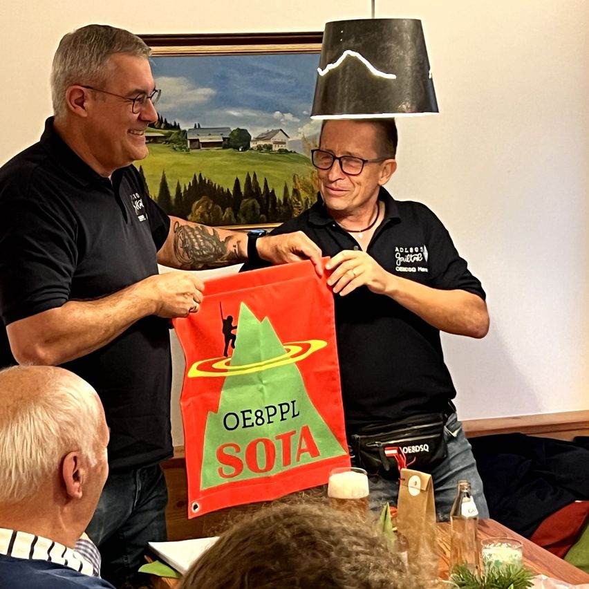 Zwei Männer halten ein rotes Banner mit 'OE8PPL SOTA' darauf. Sie lächeln und halten das Banner über einem Tisch. Ein Gemälde hängt an der Wand hinter ihnen.