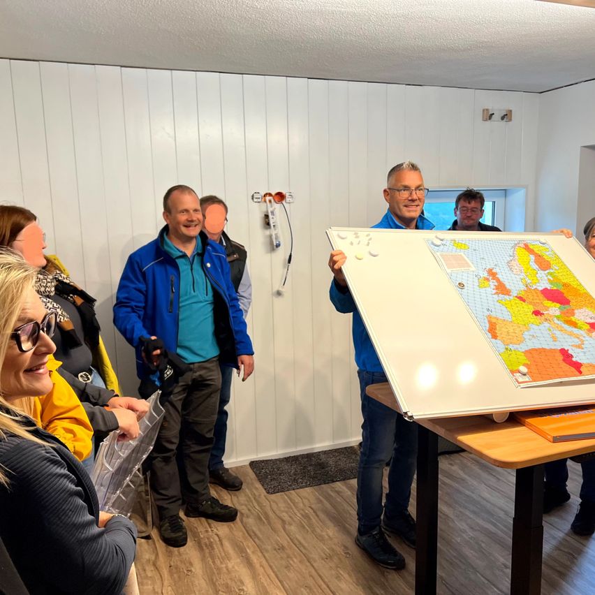 Eine Gruppe von Menschen in einem Raum mit einer großen Karte von Europa auf einer Tafel. Ein Mann hält die Tafel, während ein anderer Mann die Karte betrachtet.