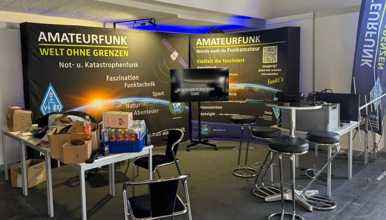 Ein Raum zeigt ein Banner mit 'Amateurfunk' und 'Surfunk'. Ein Monitor und ein QR-Code sind auf dem Banner. Flaschen und andere Gegenstände stehen auf einem Tisch. Stühle und ein Schreibtisch sind angeordnet. Scheinwerfer sind an der Decke montiert.