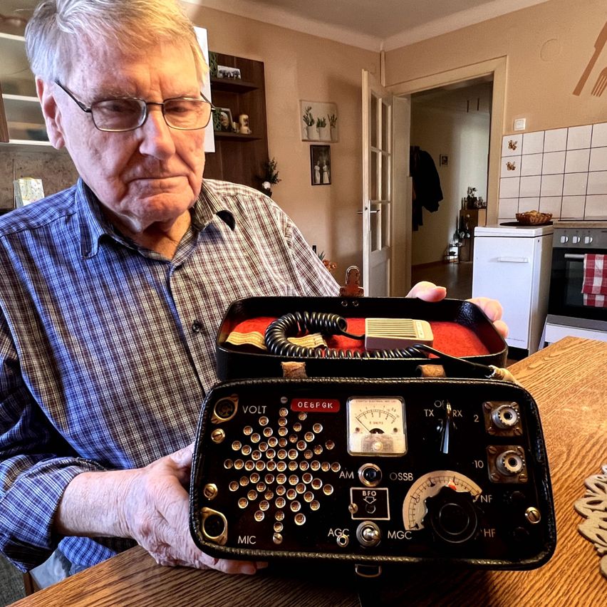Ein älterer Mann hält ein altes Radiogerät mit Mikrofon und verschiedenen Tasten und Schaltern. Er sitzt an einem Holztisch.