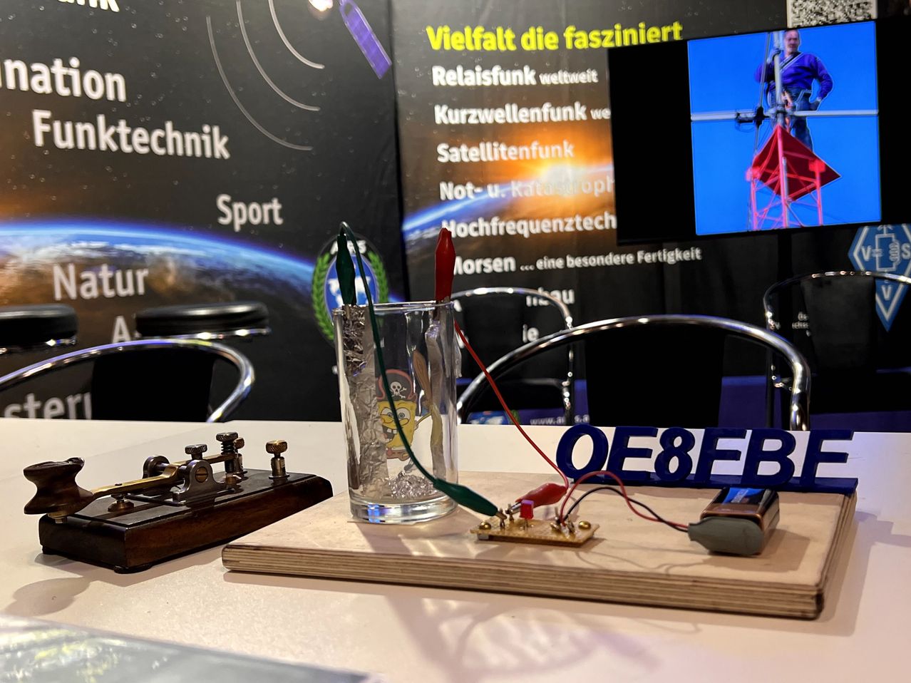 Ein Schreibtisch-Setup mit einem Glas, das Flüssigkeit enthält, Drähten und einer Leiterplatte, mit einem Morse-Code-Gerät links und einem blauen Schild mit der Aufschrift OE8EB.