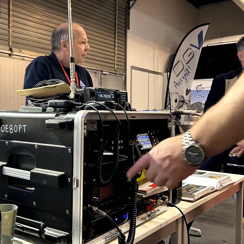 Ein Mann steht vor einer großen Funkgeräte-Anlage und zeigt darauf, während eine andere Person sitzt und ihn ansieht.