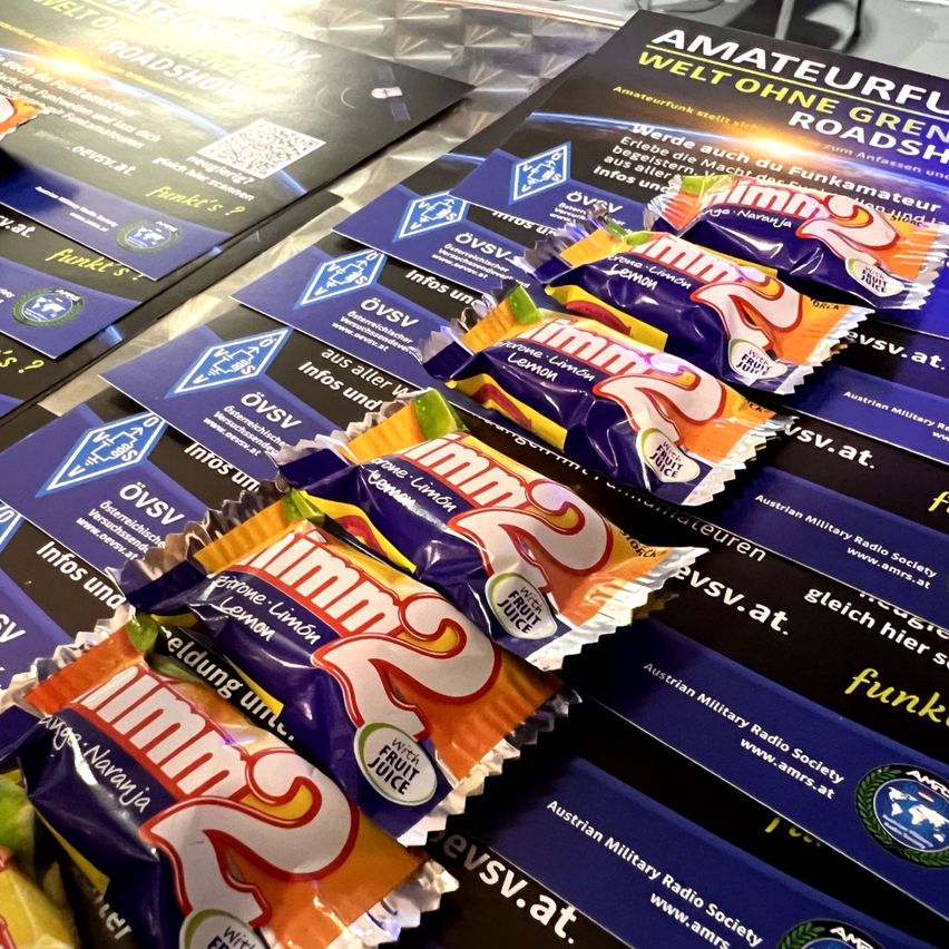 Mehrere Packungen von Süßigkeiten, in Orange und Blau verpackt, sind auf einem Tisch angeordnet. Die Packungen haben den Text 'Imms' in großen Buchstaben und enthalten Fruchtsaft. Es gibt Broschüren mit Informationen über Amateurfunk.