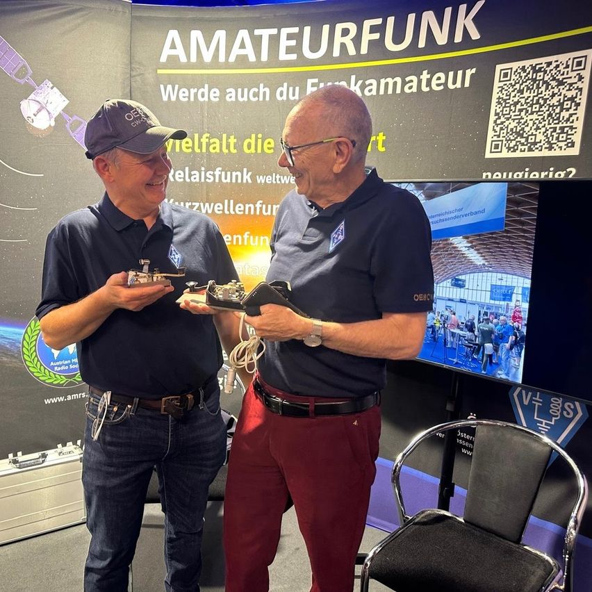 Zwei Männer in identischen blauen Poloshirts und Jeans stehen vor einem Hintergrund mit einem Satellitenbild und der Aufschrift 'Amateurfunk'. Einer hält ein kleines elektronisches Gerät, der andere ein größeres. Sie tragen beide Gürtel, einer mit einer Uhr.
