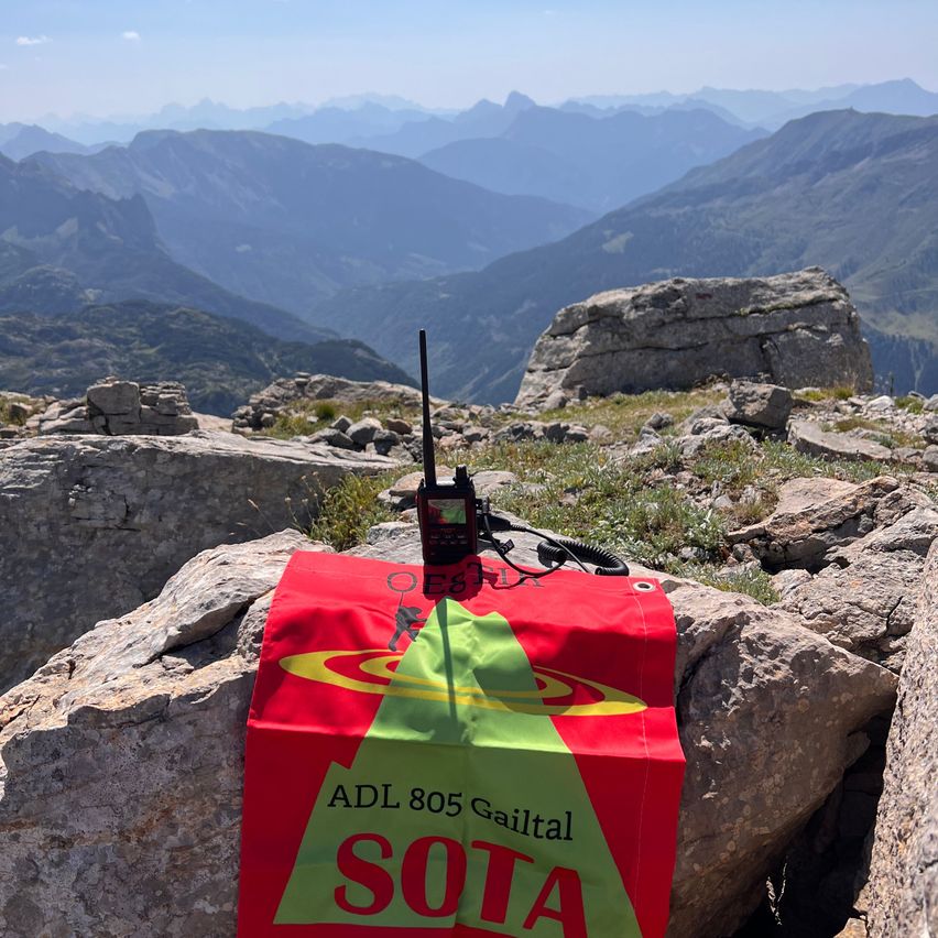 Ein rotes Banner mit dem Text 'ADL 805 Gailtal SOTA' liegt auf einem felsigen Berggipfel. Eine Funkantenne ist auf dem Banner positioniert. Der Hintergrund zeigt eine Berglandschaft mit klarem Himmel.
