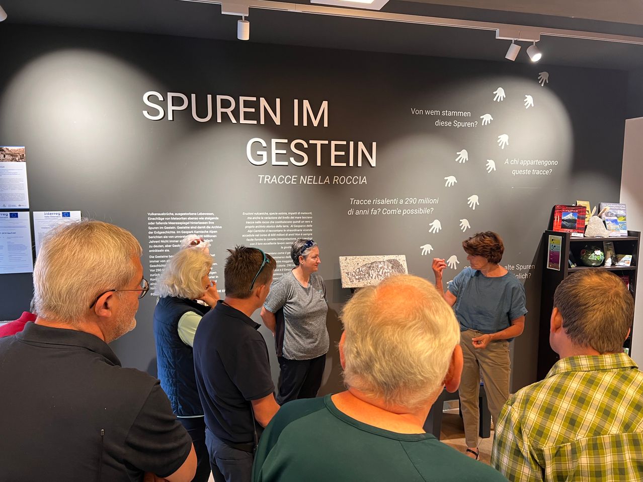 Eine Gruppe von Menschen steht vor einer Wand mit einem großen Text, der Spuren im Gestein lautet. Sie betrachten eine Wand mit Zeichnungen und Text.