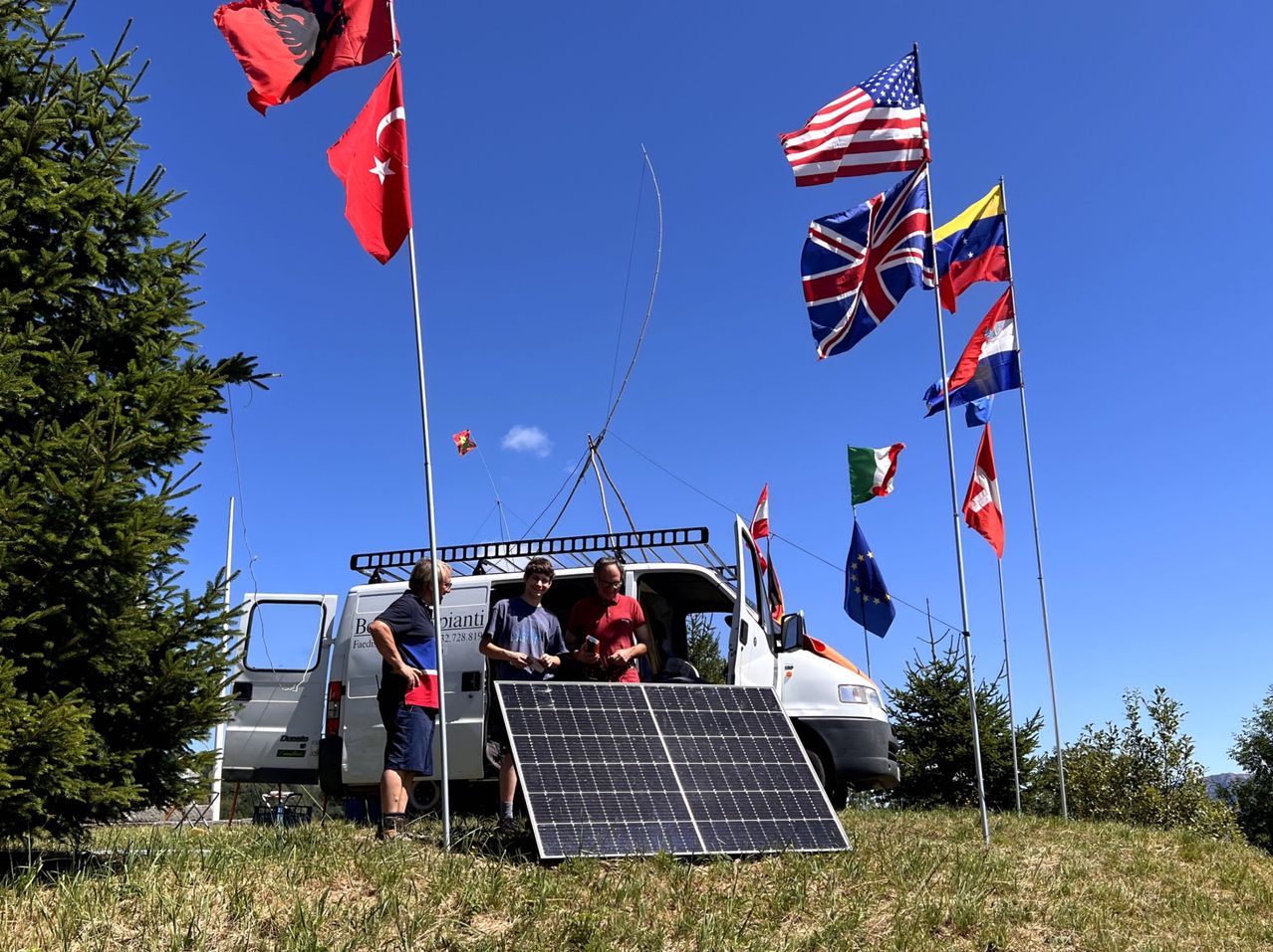 Drei Personen stehen neben einem Van mit einem Solarpanel auf dem Boden, umgeben von verschiedenen Flaggen an Stangen.