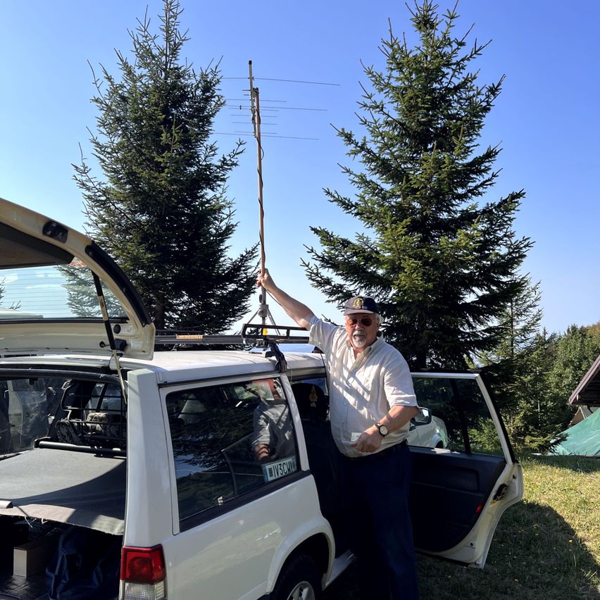 Ein Mann steht neben einem weißen SUV mit offenem Kofferraum und hält eine lange Antenne. Er trägt einen Hut und eine Sonnenbrille. Hinter ihm sind zwei Kiefern und ein klarer blauer Himmel zu sehen.