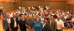 Eine Gruppe von Menschen steht in einem Konferenzraum. Einige tragen Brillen, Anzüge und Lederschuhe. Zwei Frauen in der Mitte tragen Brillen, und eine von ihnen hat eine Uhr an ihrem Handgelenk. Hinter ihnen sitzen Menschen auf Stühlen. Links stehen weiße Tische und Podiums.