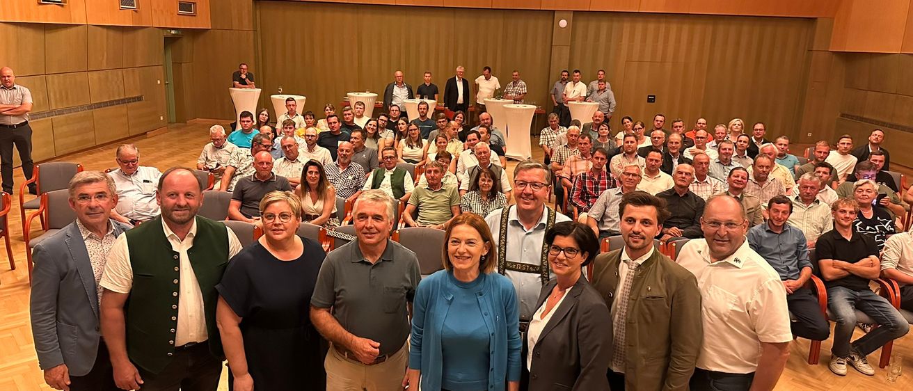 Eine Gruppe von Menschen steht in einem Konferenzraum. Einige tragen Brillen, Anzüge und Lederschuhe. Zwei Frauen in der Mitte tragen Brillen, und eine von ihnen hat eine Uhr an ihrem Handgelenk. Hinter ihnen sitzen Menschen auf Stühlen. Links stehen weiße Tische und Podiums.