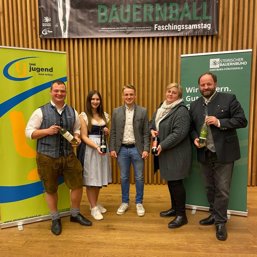 Fünf Personen stehen vor einer Holzwand und lächeln für ein Foto. Sie halten Flaschen Wein in den Händen. Die Wand hat ein grünes und weißes Banner mit dem Text 'Bauernball Faschingssamstag'.
