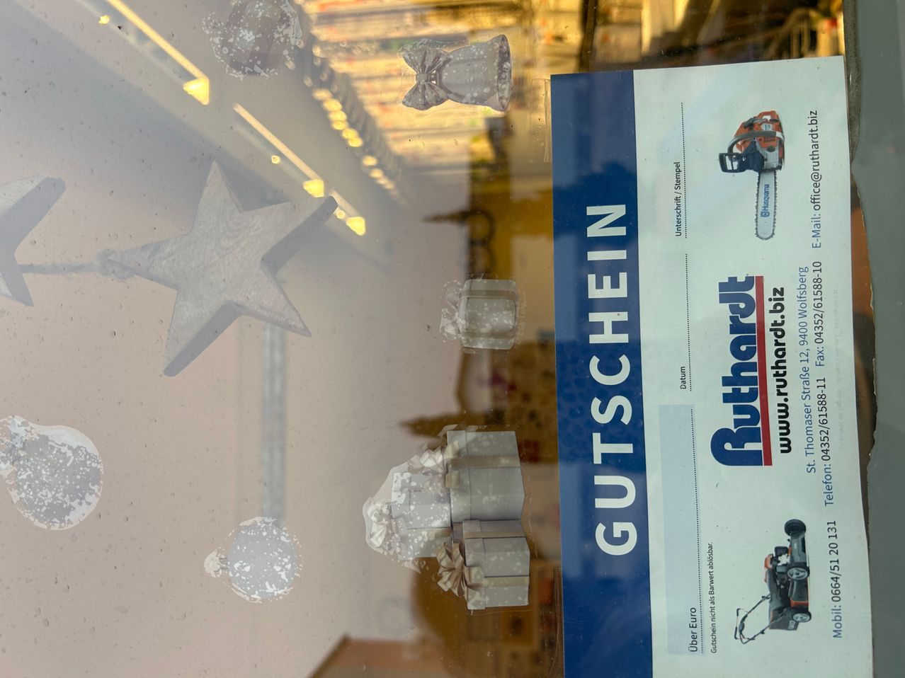 Ein Schaufenster zeigt einen Stern und einige Gegenstände. Eine Werbung für Ruthardt.biz ist sichtbar, mit einer Kettensäge. Der Hintergrund ist verschwommen, mit Reflexionen von Lichtern und Objekten.